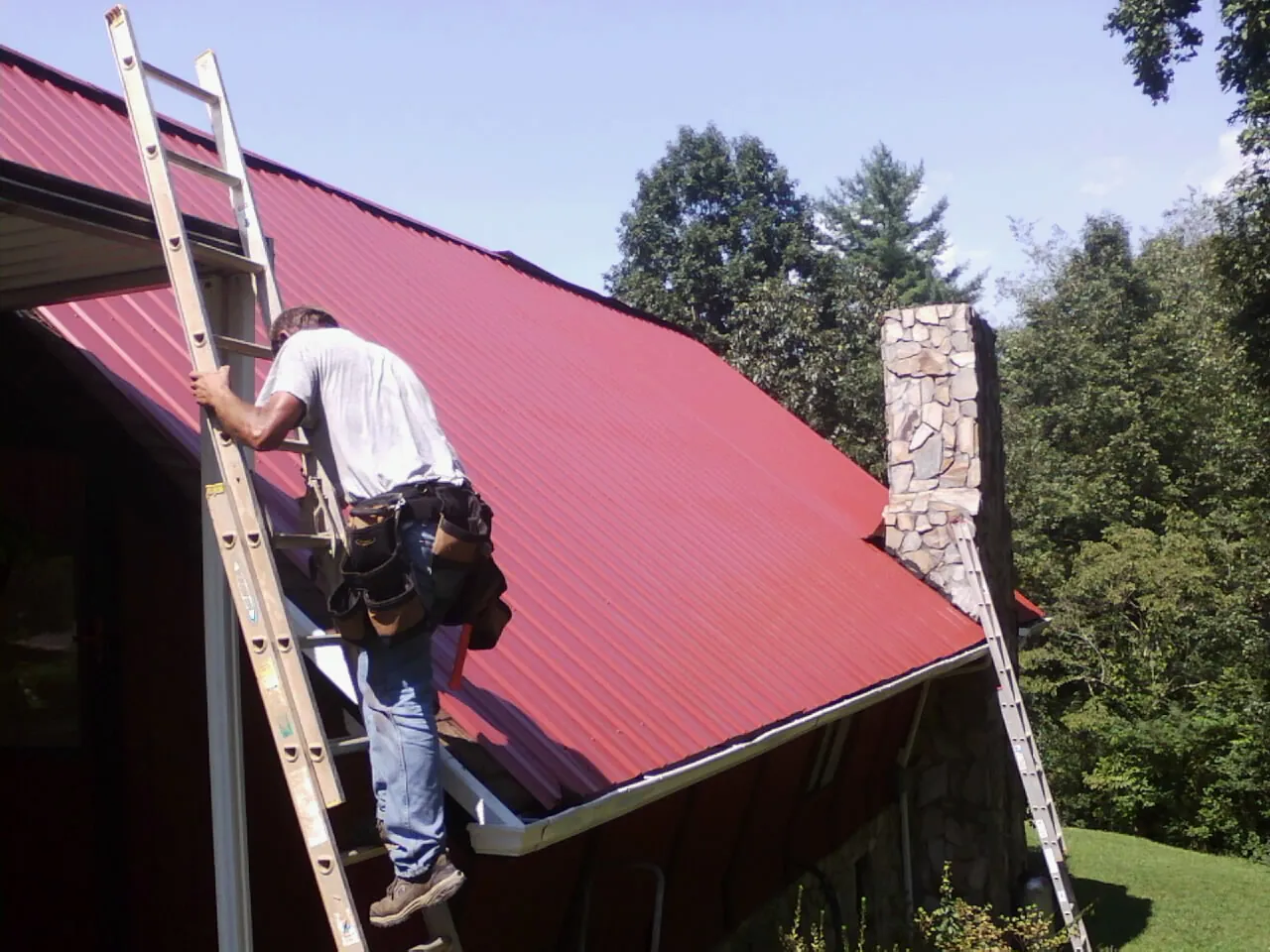 Roofer on a ladder beside a red metal roof and stone chimney — Soffit & Fascia Repair by Roofing Brothers in Vergas